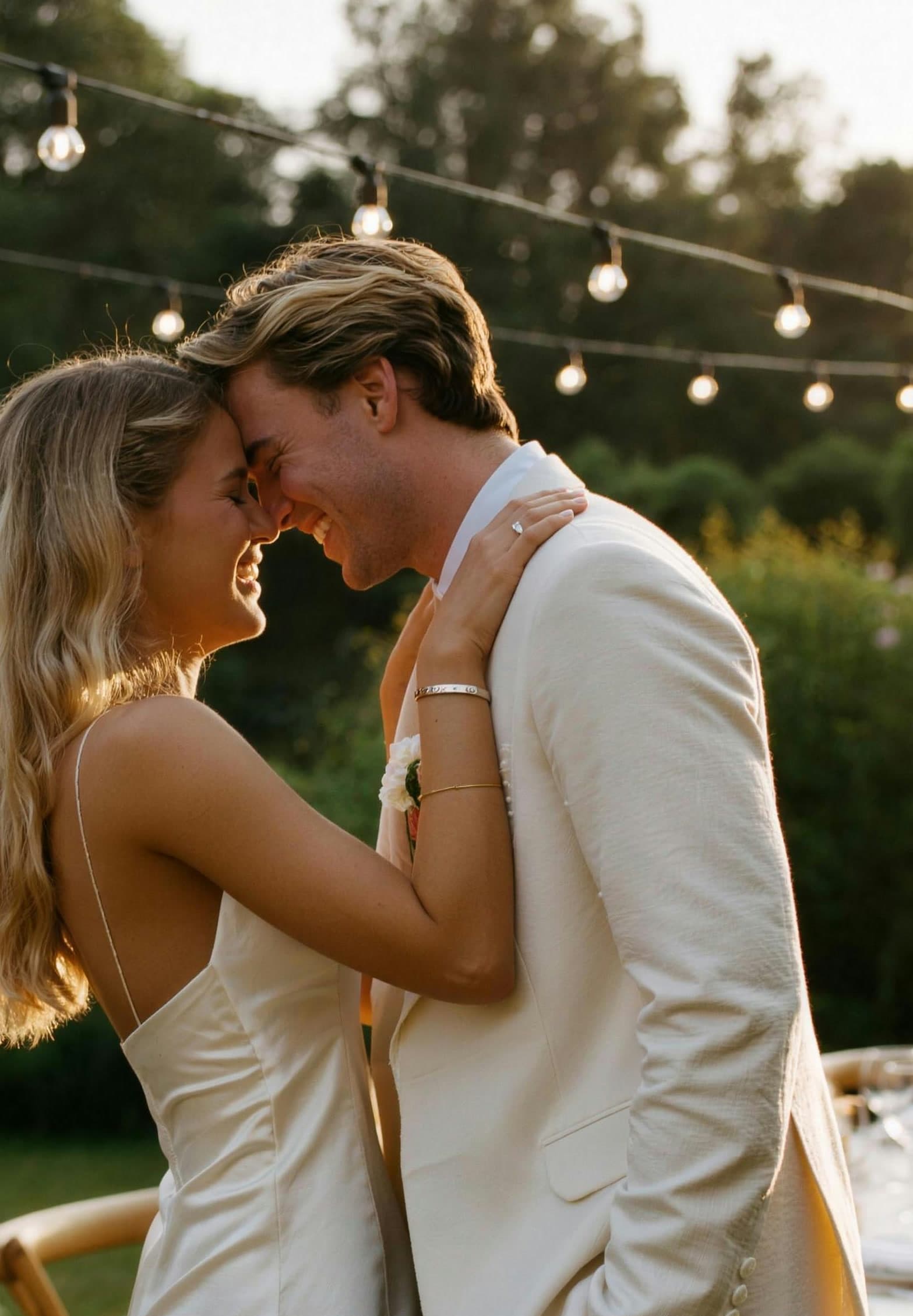 A warm golden-hour photo of the couple embracing under string lights, foreheads touching.